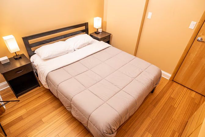 Cozy minimalist bedroom with a double bed in a beige comforter, white pillows, two bedside tables with lamps, warm hardwood floors and tan walls.