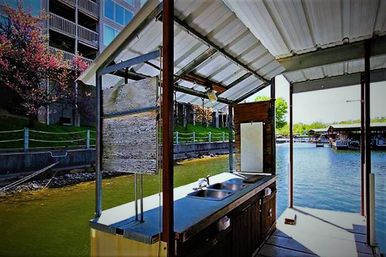 Covered lakeside dock kitchenette with stainless double sink and countertop overlooking a calm marina, shoreline trees, and boat slips.