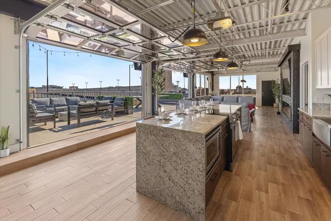 Sunlit urban loft with open-concept kitchen featuring a granite island and pendant lights, glass roll-up doors opening to a rooftop patio with lounge seating and city skyline views.