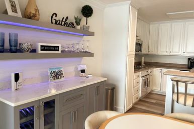 Bright open-concept home kitchen with white cabinets, gray beverage bar and wine cooler, marble-look countertop, purple LED-lit floating shelves with wine glasses and 'Gather' sign, round dining table on wood-look floors.