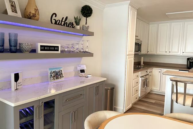 Bright open-concept home kitchen with white cabinets, gray beverage bar and wine cooler, marble-look countertop, purple LED-lit floating shelves with wine glasses and 'Gather' sign, round dining table on wood-look floors.