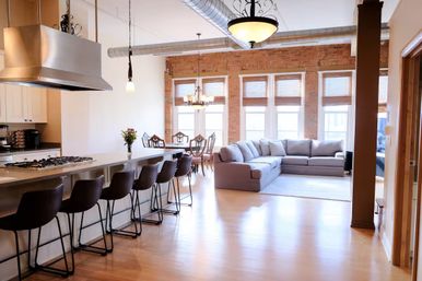 Sunlit modern loft with open-concept kitchen island and bar stools, stainless range hood, exposed brick wall, dining table, large windows and gray sectional on hardwood floors.