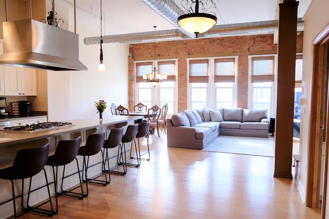Sunlit modern loft with open-concept kitchen island and bar stools, stainless range hood, exposed brick wall, dining table, large windows and gray sectional on hardwood floors.