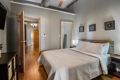Cozy loft-style urban bedroom with exposed ductwork, warm hardwood floors, queen bed with neutral quilt and white pillows, wooden headboard, geometric wall art, nightstand lamp, and open doorway to a hallway and closet.