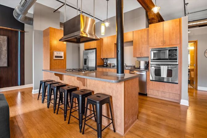Bright modern industrial loft kitchen with long island and five black metal stools, stainless steel appliances and range hood, warm maple cabinets, exposed ductwork and hardwood floors.
