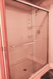 Residential bathroom sliding glass shower with white tiled walls, chrome handle and fixtures, corner shelves with shampoo bottle, overhead showerhead and gray bath mat