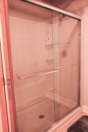 Residential bathroom sliding glass shower with white tiled walls, chrome handle and fixtures, corner shelves with shampoo bottle, overhead showerhead and gray bath mat