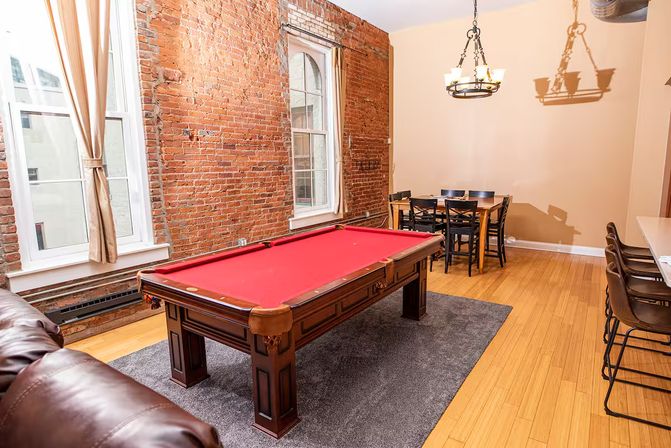 Urban loft game room with exposed brick wall and arched windows, red-felt pool table on gray rug, leather sofa, wooden dining table and bar stools under chandelier