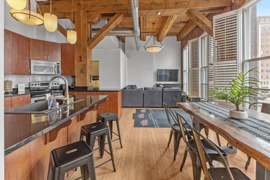 Stylish urban loft with exposed wooden beams and ductwork, open-concept kitchen featuring a dark granite island and metal bar stools, wooden dining table with chairs and potted plant, bright living area with gray sofa and wall-mounted TV, and tall city windows with shutters.