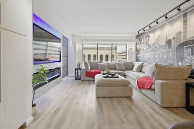 Sunlit downtown apartment living room with oversized light-gray sectional and red throws, light wood floors, subway mural wall, large window overlooking nearby historic building, wall-mounted TV with purple LED and built-in fireplace.