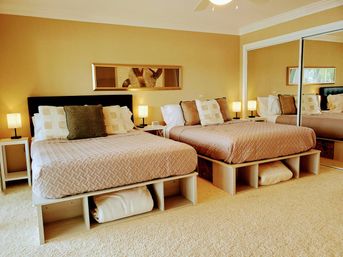 Inviting bright bedroom with two platform beds dressed in beige quilted bedding, patterned throw pillows, matching nightstands and lamps, mirrored closet doors and under-bed storage on plush carpet.