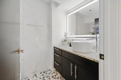 Sleek modern apartment bathroom with round white vessel sink on speckled granite countertop, dark wood vanity with stainless handles, LED backlit mirror, marble shower walls and pebble-stone floor.