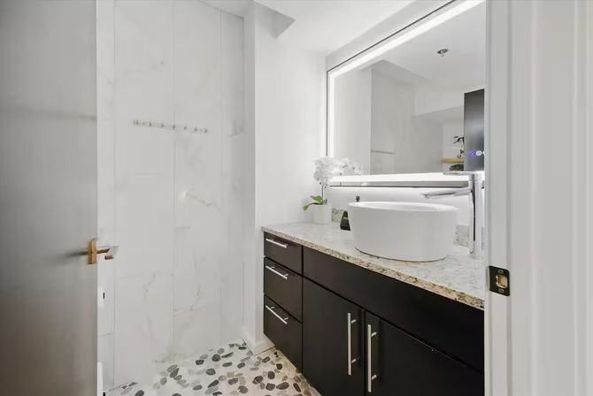 Sleek modern apartment bathroom with round white vessel sink on speckled granite countertop, dark wood vanity with stainless handles, LED backlit mirror, marble shower walls and pebble-stone floor.