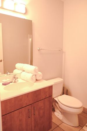 Compact neutral bathroom with wood vanity and white sink, chrome faucet, stacked rolled white towels on the countertop, wall mirror and light fixture, white toilet and tiled floor.