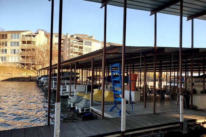 Lakefront marina with covered boat slips, two yellow-covered jet skis, a bright blue boarding ladder on the dock, and sunlit waterfront condominiums in the background.