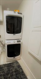 Cozy laundry nook with stacked white front-load washer and dryer, gray shag rug, and white cabinetry in a compact utility room.
