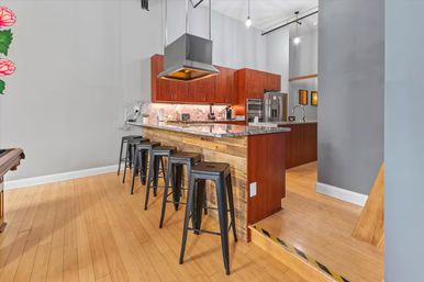 Open-concept modern-industrial kitchen with reclaimed wood breakfast bar, granite countertop, five black metal bar stools, stainless steel range hood and appliances, pendant lights, gray walls and warm hardwood floors.