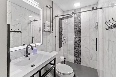 Sleek modern bathroom with white marble-look tile, illuminated LED mirror, black faucet and hardware, glass sliding shower with mosaic accent strip and rainfall shower panel, wall-mounted towel rack and white toilet.