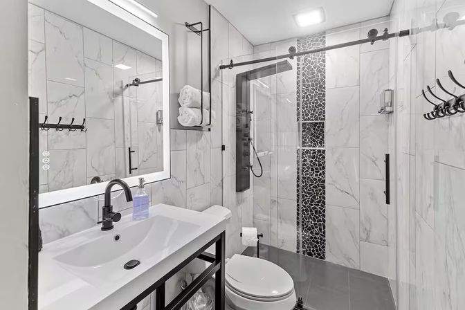Sleek modern bathroom with white marble-look tile, illuminated LED mirror, black faucet and hardware, glass sliding shower with mosaic accent strip and rainfall shower panel, wall-mounted towel rack and white toilet.