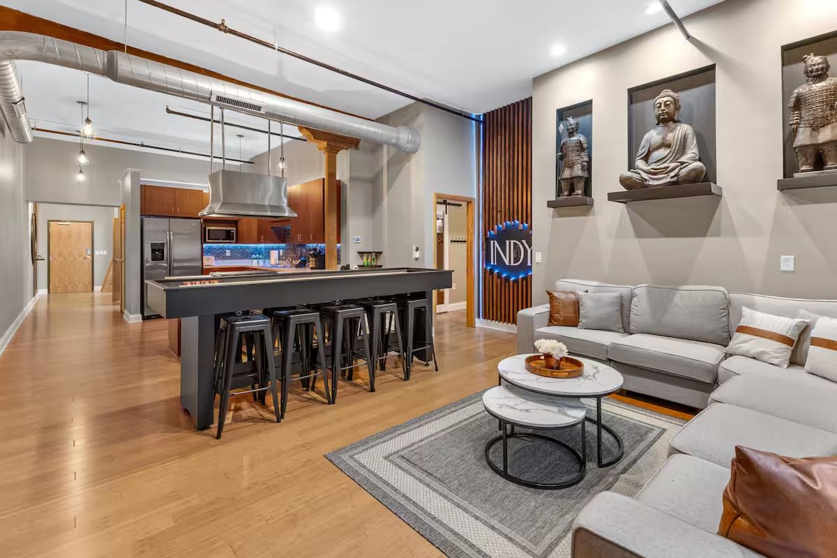 Industrial-chic open-concept loft with a long black kitchen island and bar stools, stainless steel hood, exposed ductwork, hardwood floors, cozy gray sectional, marble nesting tables and Buddha wall sculptures.