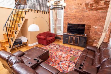 Cozy urban loft living room with exposed brick wall, hardwood floors, brown leather sectional with cup holders, colorful patterned area rug, red loveseat, flat-screen TV on a rustic media console, and wooden staircase with wrought-iron railing.