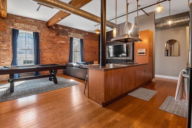 Industrial urban loft with open-concept kitchen and living area — exposed brick walls, hardwood floors, arched windows, pool table, wood beams, and stainless-steel range hood.