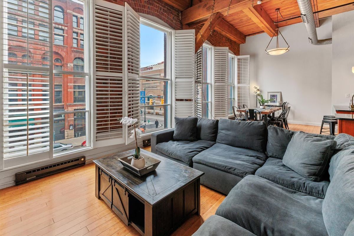 Sunlit urban loft living room with dark gray sectional sofa, wooden coffee table and hardwood floors, exposed brick and timber beams, tall windows with white plantation shutters overlooking a downtown street and dining area in the background.