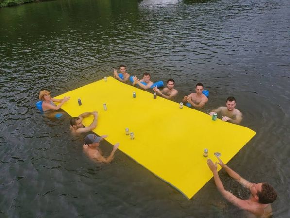 Group of adults in a lake holding a large bright yellow floating table mat with canned drinks on top — summer outdoor floating party scene.
