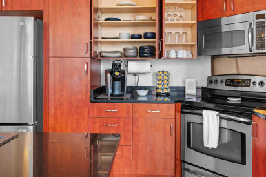 Modern kitchen with cherry wood cabinets, stainless steel refrigerator, microwave and oven, black granite countertops, open shelving with plates and glasses, and a coffee pod machine on the counter.