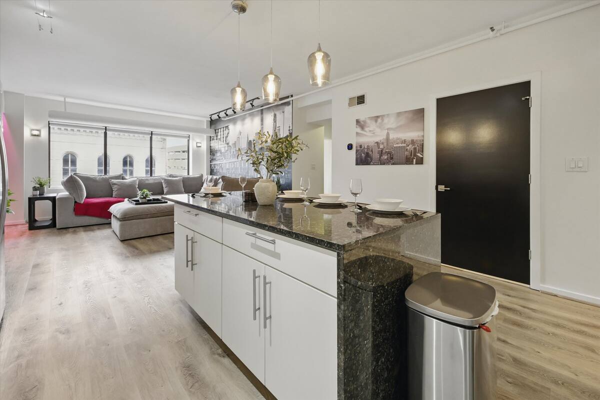Bright open-plan urban apartment interior featuring a dark granite kitchen island with white cabinets and pendant lights, light wood floors, stainless trash can, gray sectional sofa with red throw by large city-view windows, and cityscape wall art.