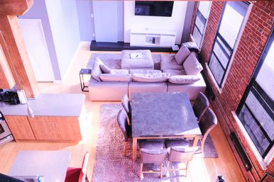 Bright industrial loft living space with exposed brick and tall windows, gray sectional sofa, square dining table with six chairs on a plush rug, open-plan kitchen island and hardwood floors.