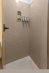 Beige corner shower stall with recessed shampoo niche, three wall-mounted soap dispensers, overhead light, and glass door handle.