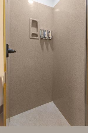 Beige corner shower stall with recessed shampoo niche, three wall-mounted soap dispensers, overhead light, and glass door handle.