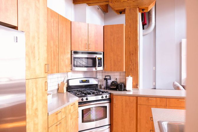 Loft-style kitchen with warm wood cabinets, stainless steel refrigerator, microwave and gas range, light countertops, white subway tile backsplash, exposed wooden beam and small countertop appliances.