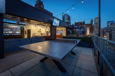 Rooftop patio at dusk with a sleek ping-pong table, outdoor kitchen and lounge under string lights overlooking an urban skyline.