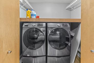 Compact home laundry closet with matching front-loading washer and dryer in metallic finish on pedestals, wooden sliding doors, wire shelf above holding detergent and cleaning supplies.