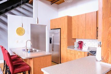 Loft-style modern kitchen with light wood cabinets, stainless steel refrigerator, quartz island with sink and chrome faucet, three red velvet bar stools, pendant lights, white subway tile backsplash and a red single-serve coffee maker for a pop of color.