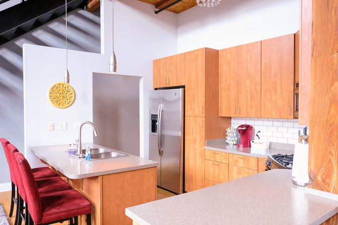 Loft-style modern kitchen with light wood cabinets, stainless steel refrigerator, quartz island with sink and chrome faucet, three red velvet bar stools, pendant lights, white subway tile backsplash and a red single-serve coffee maker for a pop of color.