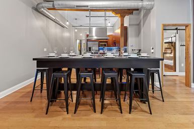 Modern industrial loft dining area with a long black communal table set with white dishware and wine glasses, black metal stools, hardwood floors, exposed ductwork, pendant lights, stainless steel kitchen and a bunk bed visible through a doorway.