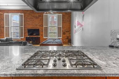 Open-concept urban loft interior with stainless steel five-burner gas cooktop on a granite kitchen island, exposed brick wall, tall arched windows with shutters, sofa, TV and a pool table in the living area.