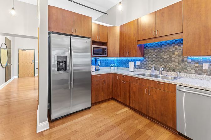 Modern apartment kitchen with stainless steel side-by-side refrigerator and dishwasher, warm wood cabinets, quartz counters, mosaic tile backsplash lit by blue LED under-cabinet lighting, double sink and hardwood floors.