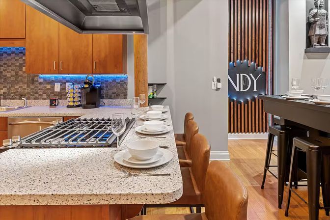 Modern open-concept kitchen with speckled quartz island and built-in gas cooktop, set with white dishes and wine glasses, brown leather bar stools, wooden cabinets and blue LED undercabinet lighting.