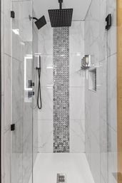 Modern walk-in shower with glass door, white marble-look tiles and vertical gray mosaic accent strip, matte black rainfall and handheld showerheads, recessed niche and wall-mounted soap dispensers