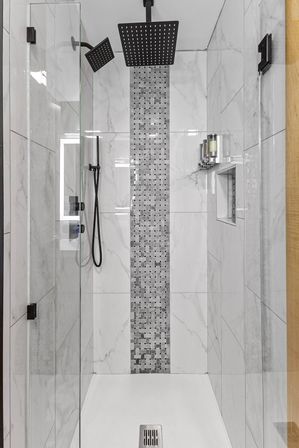Modern walk-in shower with glass door, white marble-look tiles and vertical gray mosaic accent strip, matte black rainfall and handheld showerheads, recessed niche and wall-mounted soap dispensers