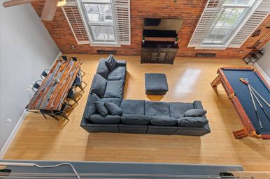 Aerial view of an open-concept urban loft with a large L-shaped gray sectional and ottoman, rustic wooden dining table with metal chairs, a pool table, exposed brick wall, tall shuttered windows, and light hardwood floors.