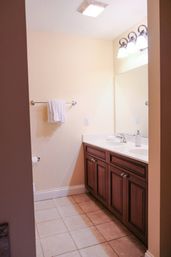 Bright residential bathroom interior with beige walls and tiled floor, dark wood double-sink vanity with white countertop, large mirror, chrome faucet, towel on wall bar and triple vanity lights.