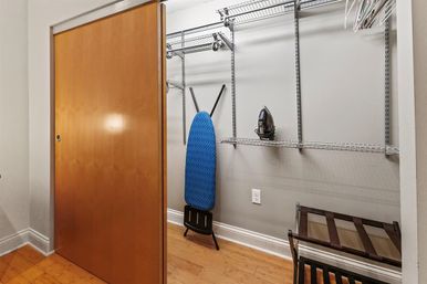 Small interior closet with a wooden sliding door, metal wire shelving, blue ironing board, iron on a shelf, folding luggage rack and hardwood floor.