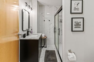 Modern compact bathroom with black vanity and marble countertop, hexagon tile floor, glass-enclosed marble shower, black fixtures, small wooden stool and framed humorous wall art.