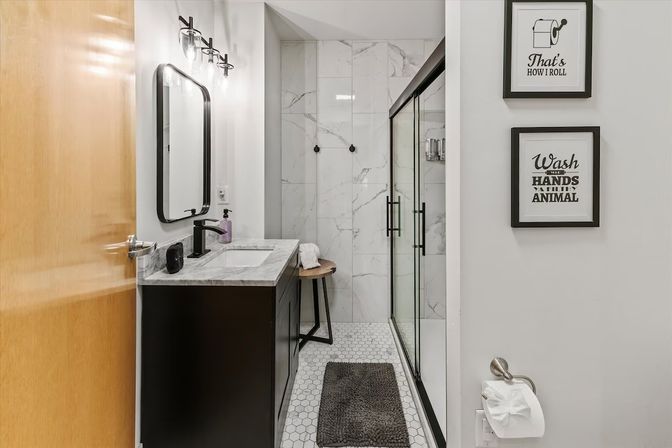 Modern compact bathroom with black vanity and marble countertop, hexagon tile floor, glass-enclosed marble shower, black fixtures, small wooden stool and framed humorous wall art.