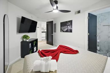 Bright modern apartment bedroom with neutral bedding, bold red knit throw and folded towels on the bed, wall-mounted TV, black ceiling fan, small console with a potted plant, and open doors leading to a kitchen and marble-tiled en-suite bathroom.